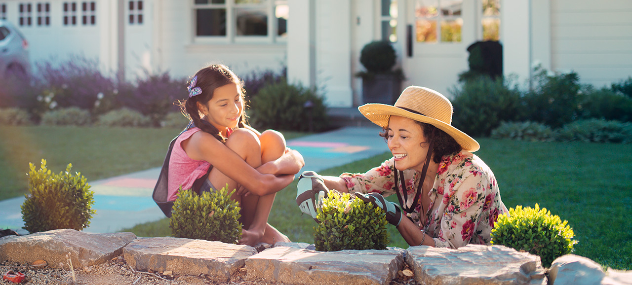 A woman trims a small shrub in her front yard with garden clippers while her granddaughter watches. yard maintenance